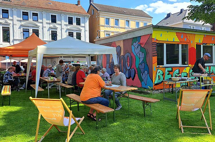 Menschen sitzen auf Bänken vor einem bunt bemalten Container.