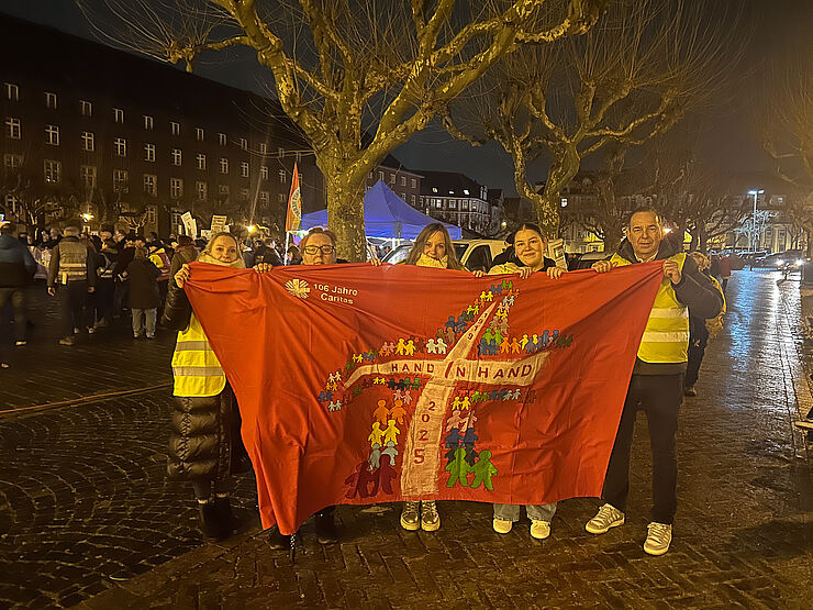 Fünf Menschen halten eine rote Fahne mit Aufdruck Hand in Hand.