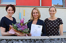 Drei Frauen stehen mit einem Blumenstrauß und einem Dokument in der Hand zusammen.